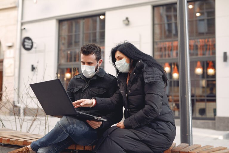 People in a masks sitting in a city with a laptop